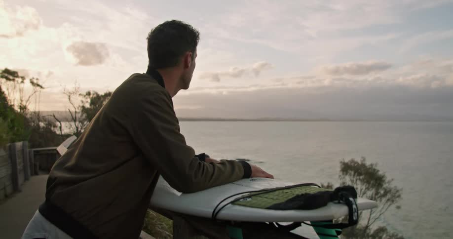 A man with a surfboard is standing on an elevated walkway looking out over a coastal area in the evening