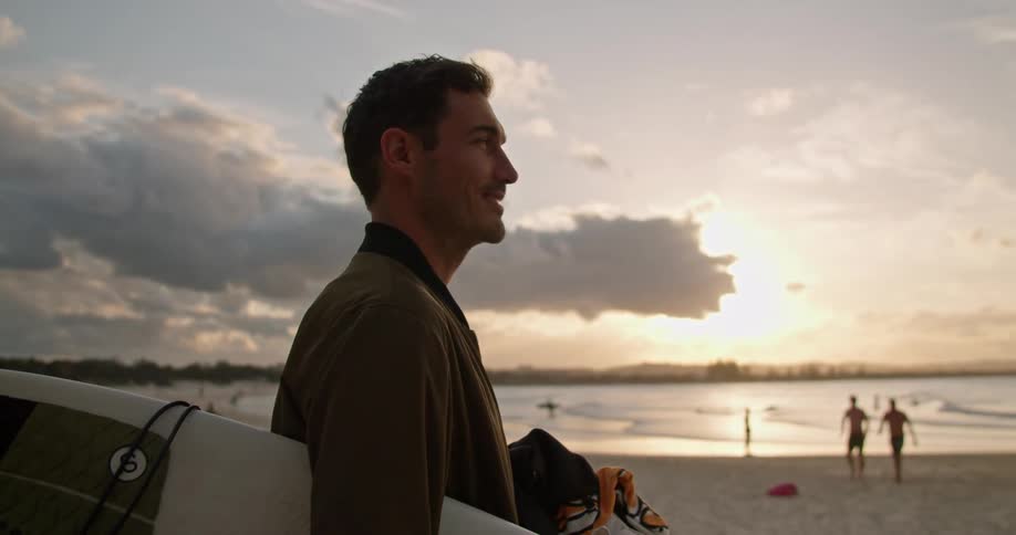 A man standing on the beach holding his surfboard and a towel looking out at the ocean