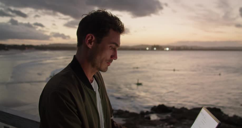 A man stands on a balcony on the shore and types on a laptop