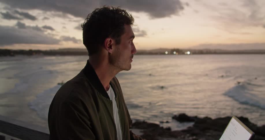 A man is standing near a railing overlooking the water and typing on a laptop computer