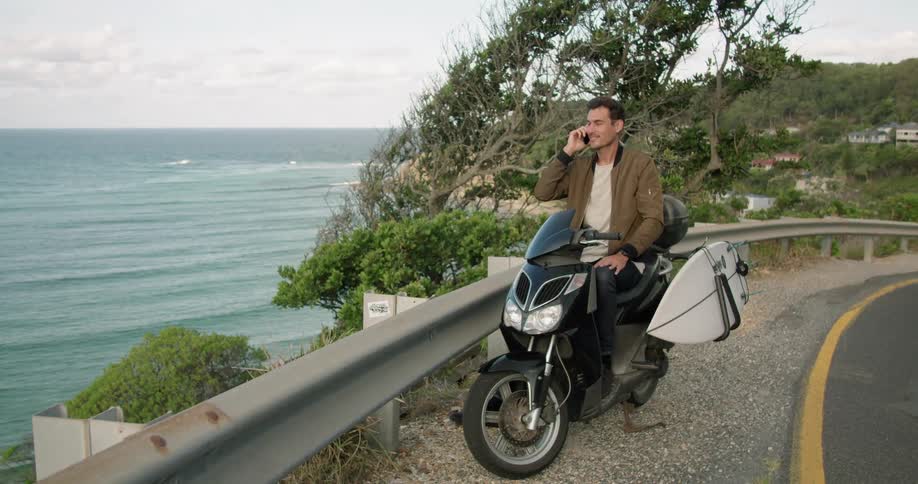 A man sits on his parked motorcycle above the shore and talks on his cell phone