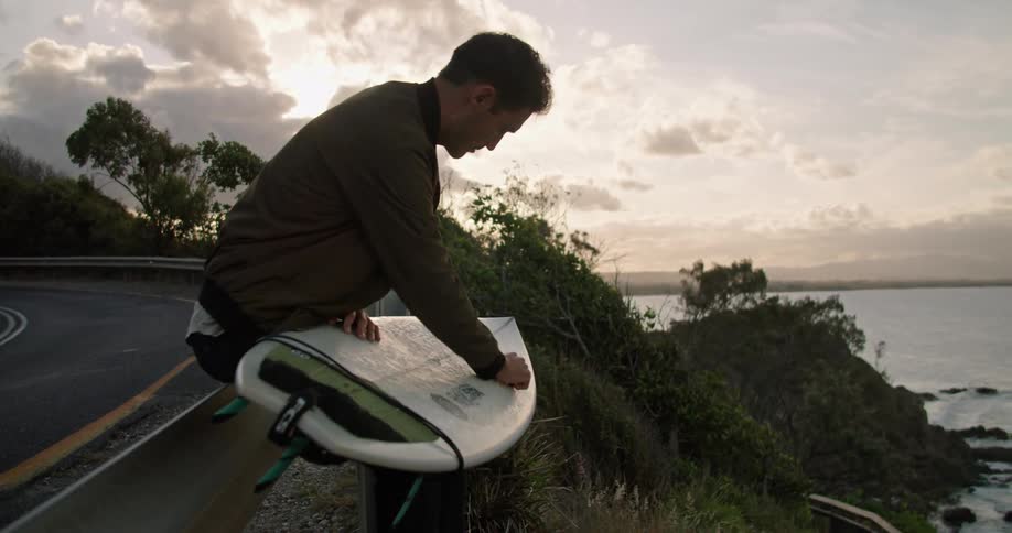 A man crouches on the ground to wax his surfboard as he stands on a cliff over the ocean