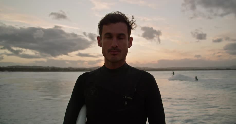 A surfer in a wet suit stares into the camera while standing on a beach in the evening