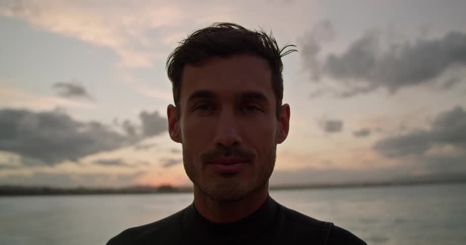 Man with wetsuit faces the camera with eye contact on the beach next to sea at dusk
