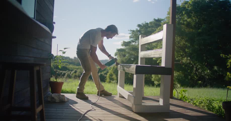 A man is adding the finishing touches to a wooden bench on a deck attached to a home in a rural area on a sunny day