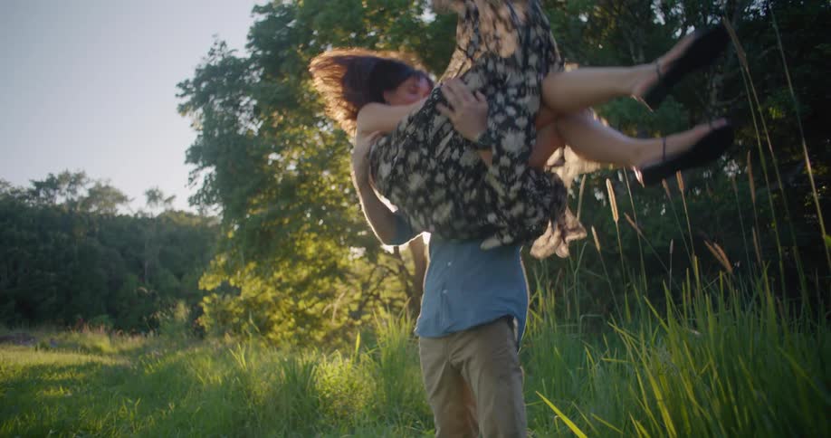 Woman in a dress gets carried on his boyfriend arms in a field at sunset