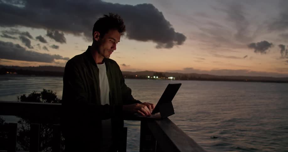 A man stands on a balcony on the shore and types on a laptop