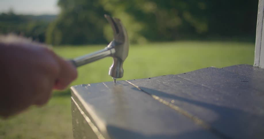 Man with tattoo hammers nail into wooden surface with field in background