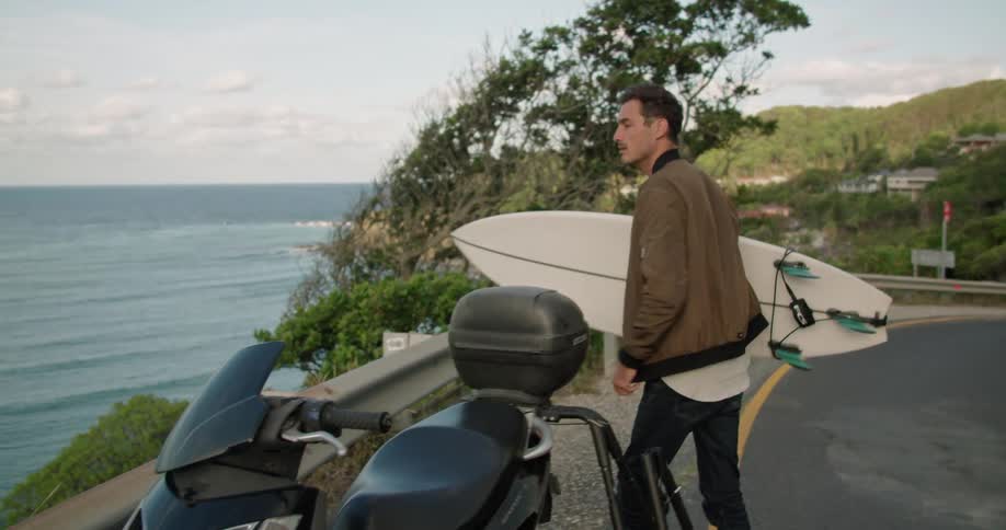A man takes a surfboard off the side of his motorcycle and walks towards the shore
