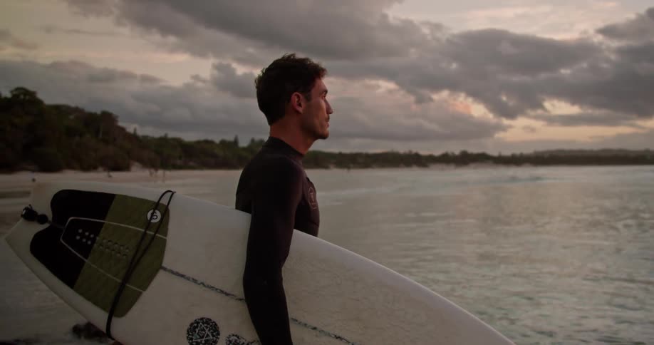 Man with wetsuit holds surfboard and looks around on the beach next to sea at dusk