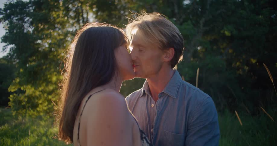 Blonde man with blue shirt kisses woman with brown wavy hair and dress lovingly looking into each others eyes in the countryside next to trees