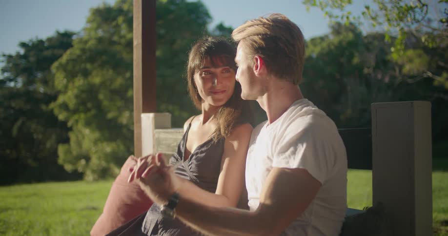 A man and a woman are sitting on a bench outside holding hands and he kisses her hand