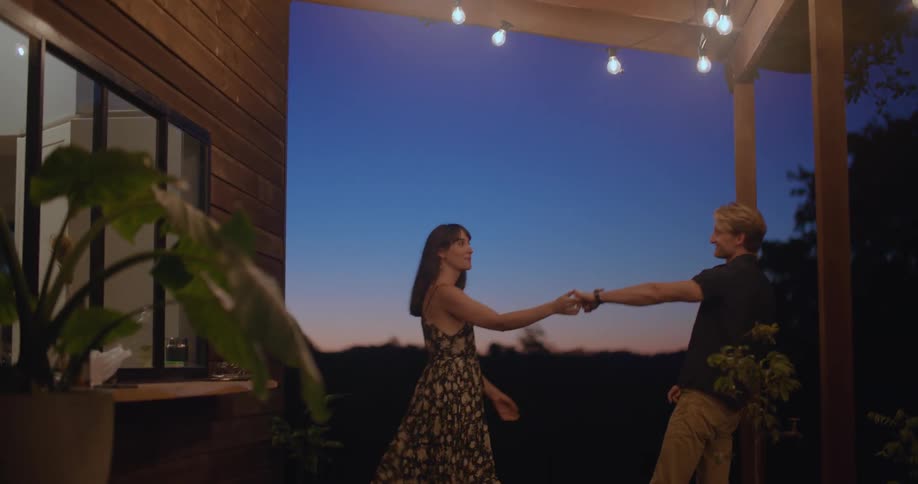 A man kisses a woman on the cheek as they dance together on the porch of a house at night