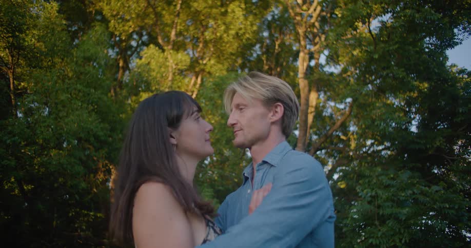 A man and a woman are standing outside near trees hugging and kissing