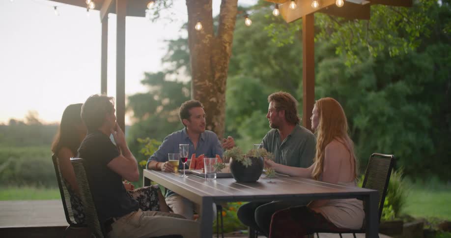 A group of friends sitting at table and talking with each other on terrace in countryside at dusk