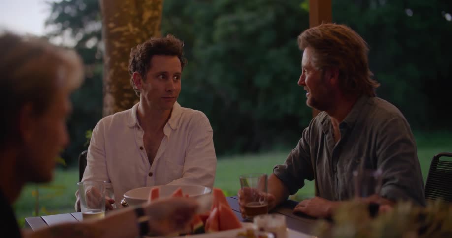 Three men are sitting at a table outdoors in a yard having conversation drinks and appetizers in the evening