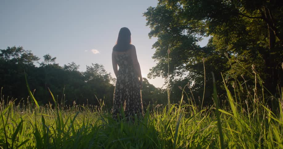 A woman wearing a sundress is standing outside in tall grass surrounded by trees