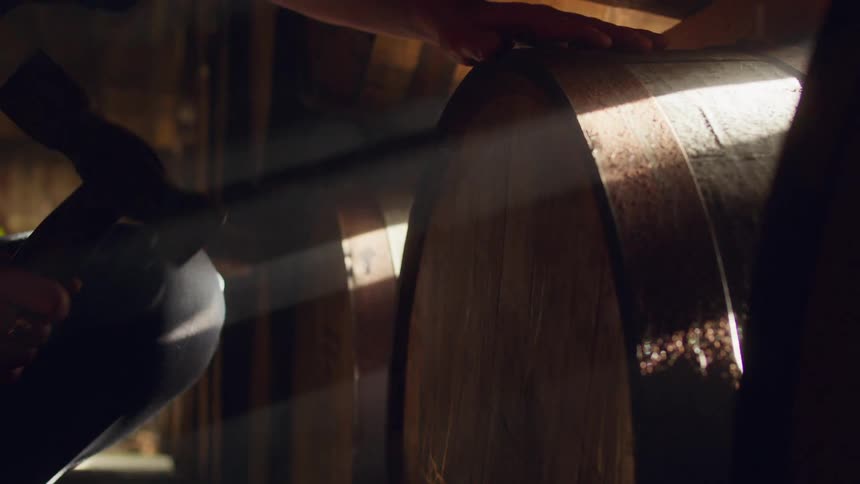 Master distiller squats in front of a large barrel in a shadowy cellar and begins tapping it with a large hammer in the daytime