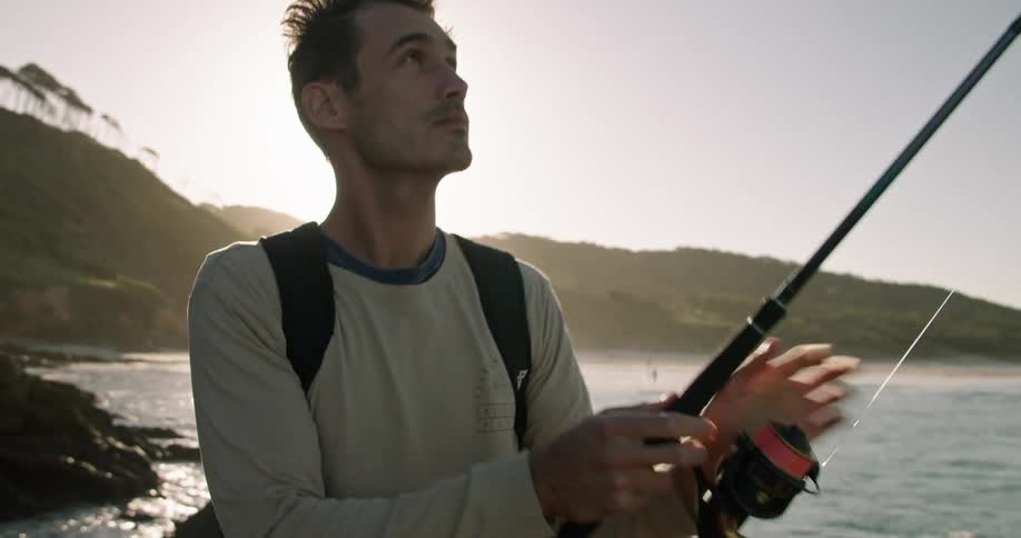 A man prepares his fishing pole while standing on a rocky coastline during the day