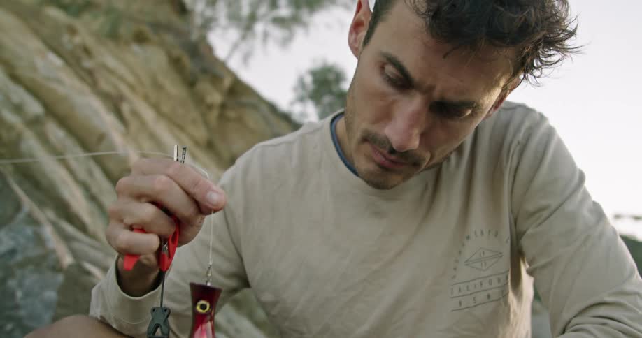 A man kneeling outdoors examines a large bait on a fish hook