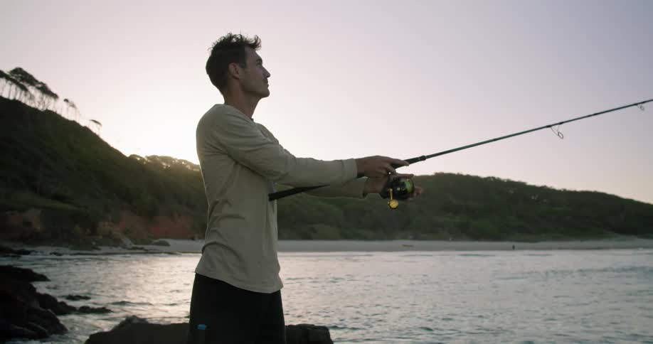 A man is standing near the water holding a fishing pole as he is casting his line