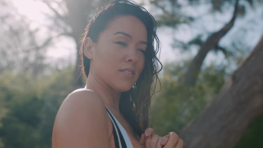 A young woman holds her hands together under a tree and looks at the ground and then releases them and looks up