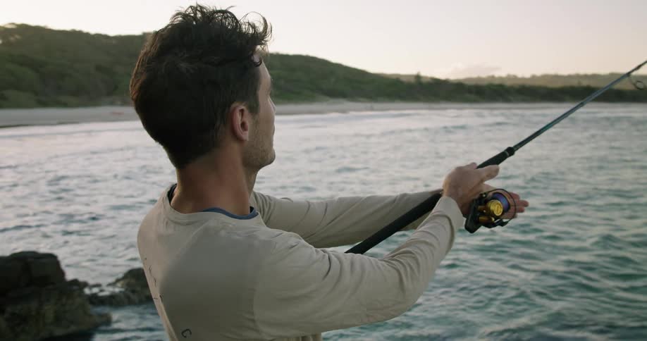 A man is standing on a rock shore while casting a fishing line into the water in the evening