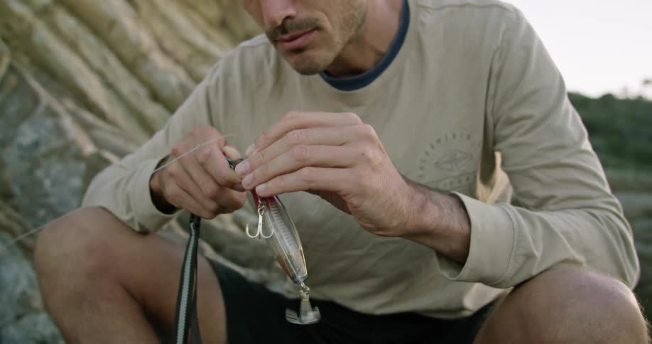 Young man sitting outdoors threading bait for fishing at sunset