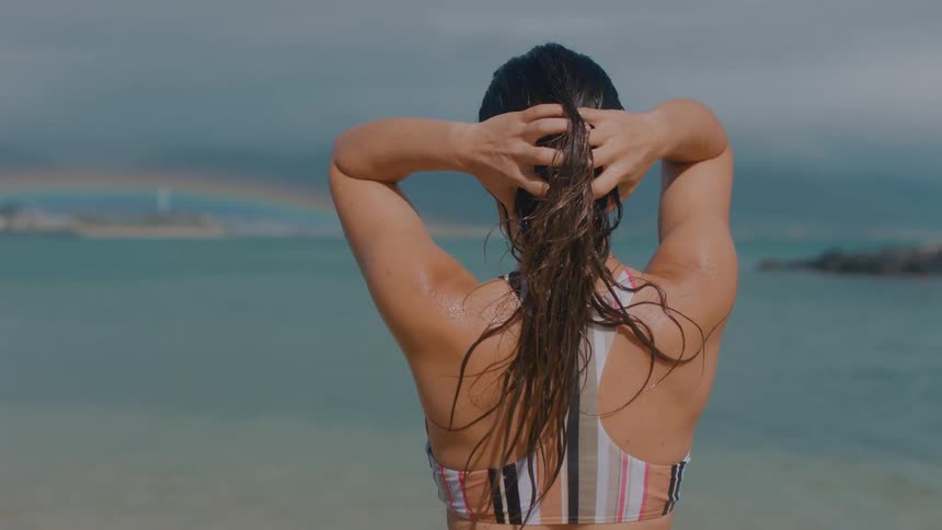 A woman standing on a beach in a swimsuit runs her hands through her hair while looking at a distant rainbow across the ocean