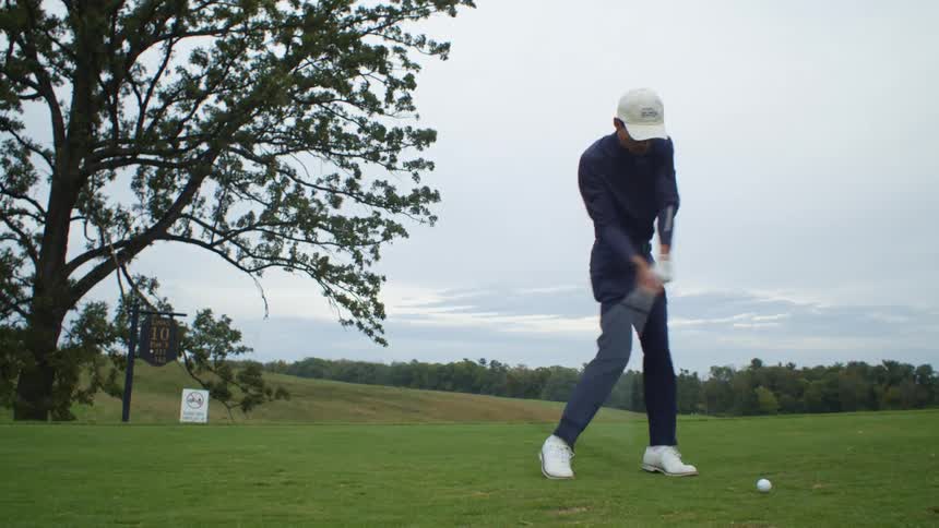 A man wearing a white hat is golfing and he swings his club hitting the ball