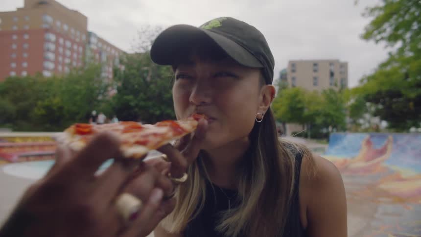 A man feeds pizza to his girlfriend as they sit outside a skate park