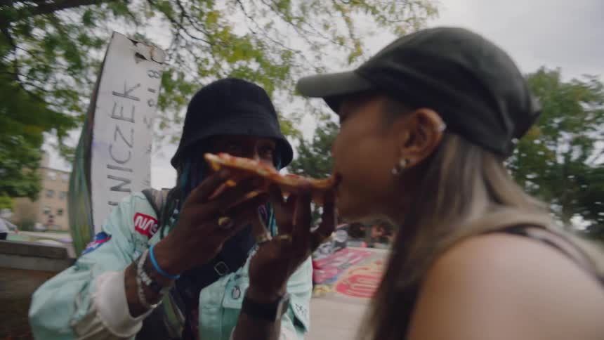 A man feeds a slice of pizza to a young woman as they sit together in a skate park on a cloudy day