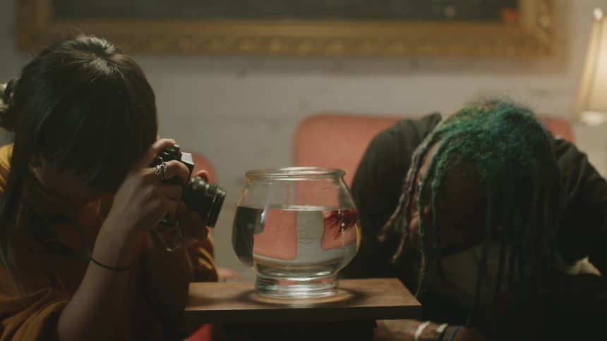 A woman is taking a photo of a fish in a fishbowl while a man looks closely at the fish while indoors