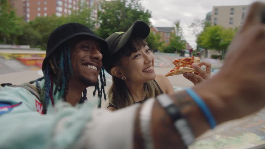 A man and woman eating pizza are taking a selfie in a skateboard park in a city on a cloudy day