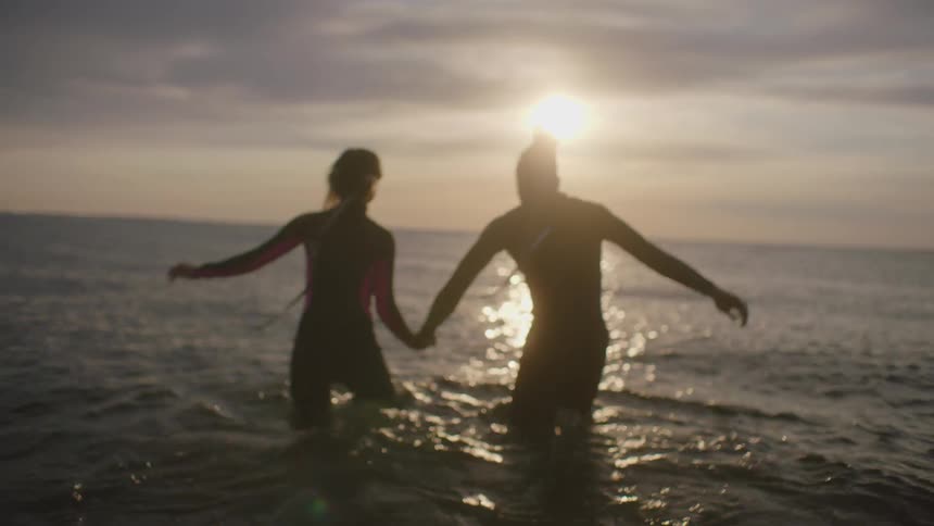 A man and woman stand on the shore and take one each others and walk in to the water together and then stop and laugh
