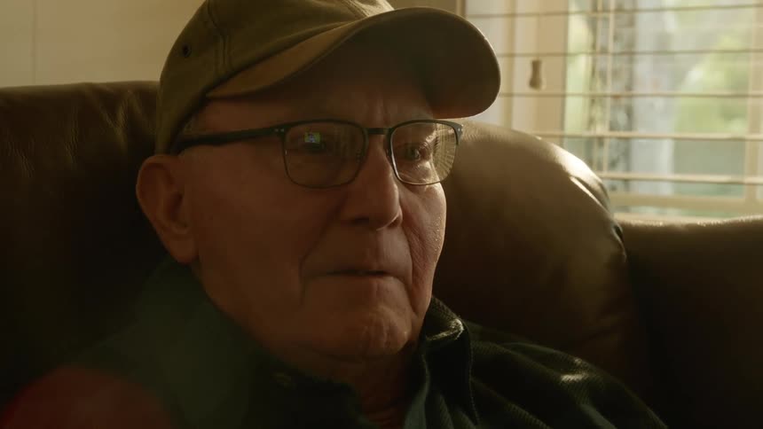Elderly man with cap and glasses sits in armchair watching television in living room at home