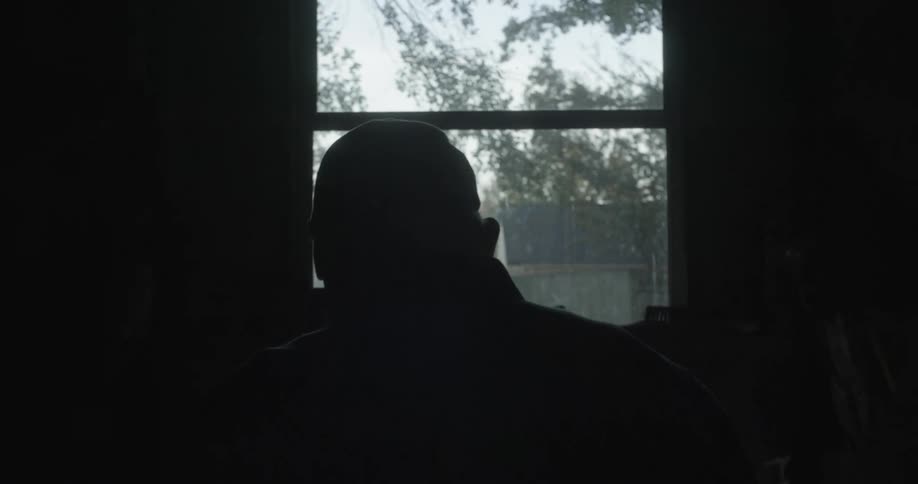 Elderly man with cap and glasses is silhouetted against the window and sky