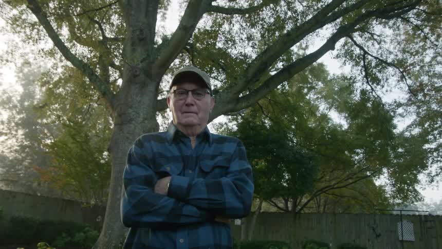 A man stands with arms crossed in a yard with a wooden fence behind him