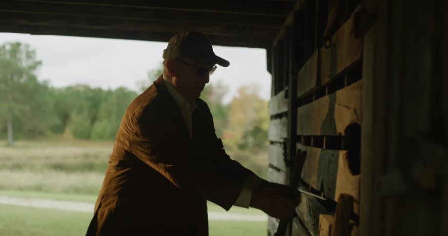 An elderly man in a barn bends down and picks up a gun and hangs it on the wall