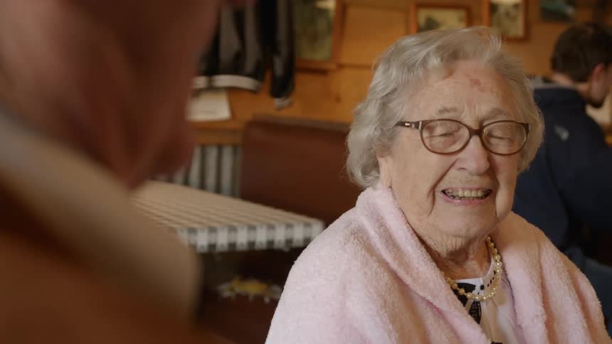 Elderly woman sits in diner smiling while talking to a man