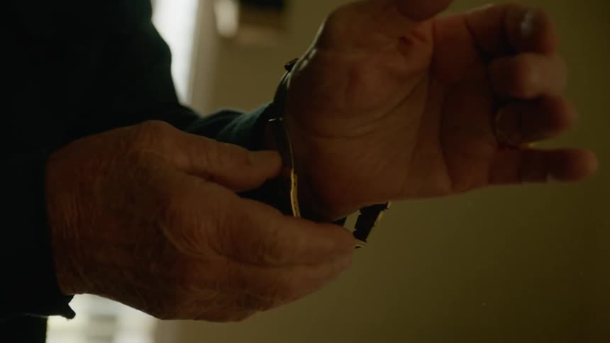 A man picks up a wristwatch from his bed and puts it on