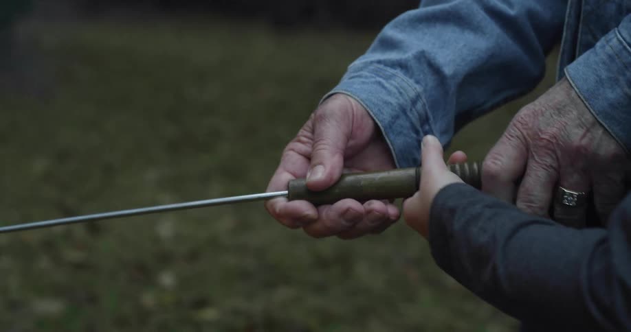 An elderly man helps a young boy hold a stick over a fire outside