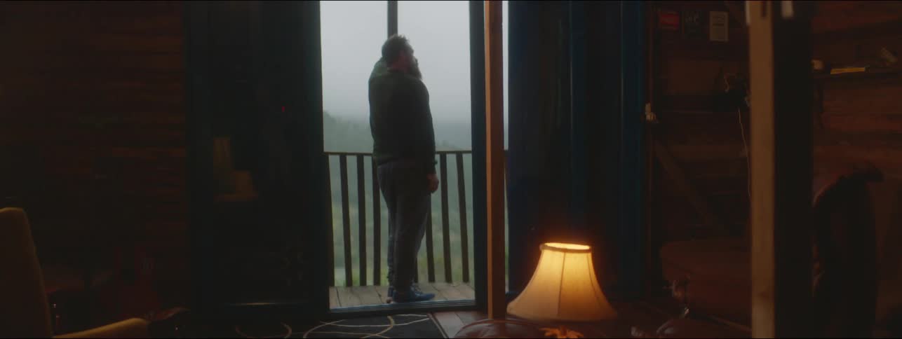 A man is walking out onto a balcony off of a cabin and leans against the railing