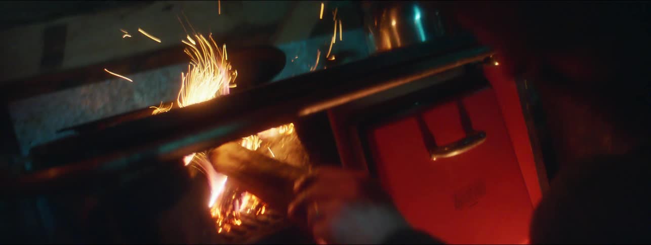 A man in a cabin pokes at a fire in a wood burning stove with a piece of wood