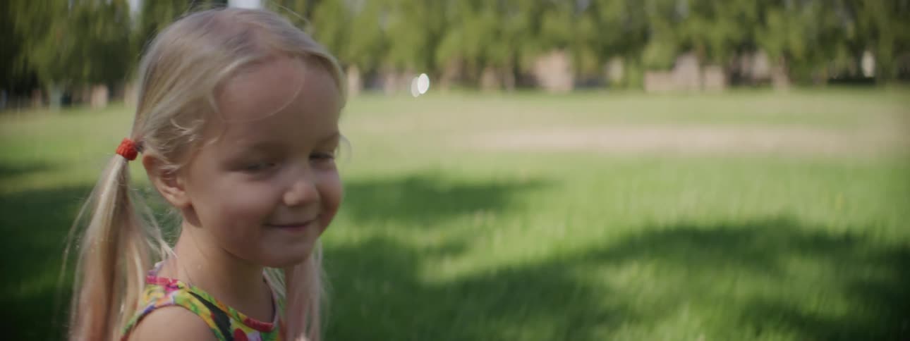 Blonde girl in a colorful dress walking on the green grass with a dandelion in her hand