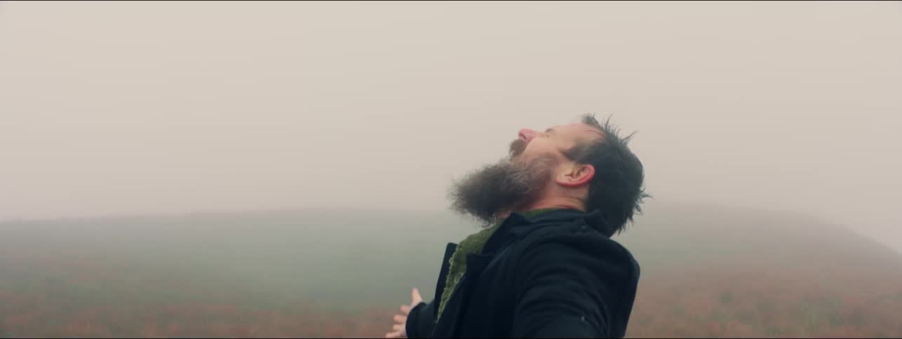 Bearded man stands near foggy hill reaching out and up