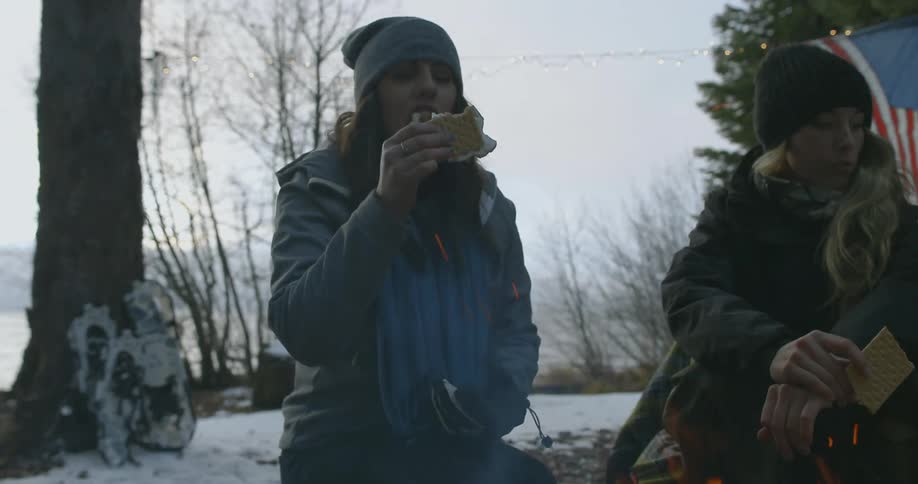 Two women sit eating and talking at a campfire