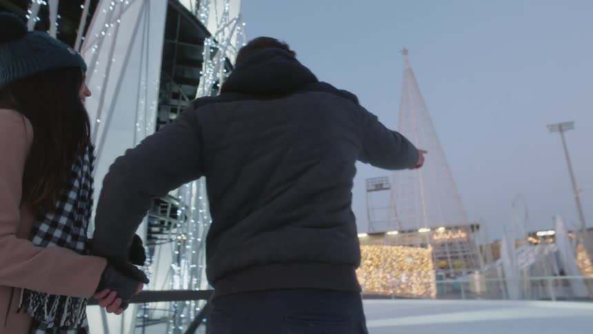 A man and woman holding hands while skating on an outdoor skating rink covered in christmas decorations