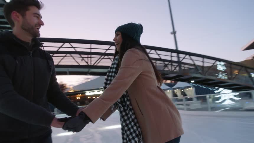Couple holding hands on an ice rink surrounded by holiday lights at night