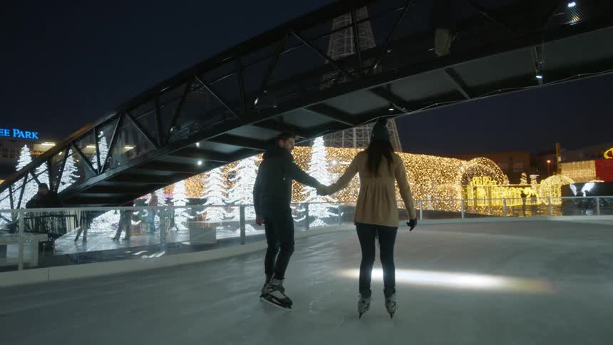 A man is teaching a woman to ice skate outside on a holiday rink at night